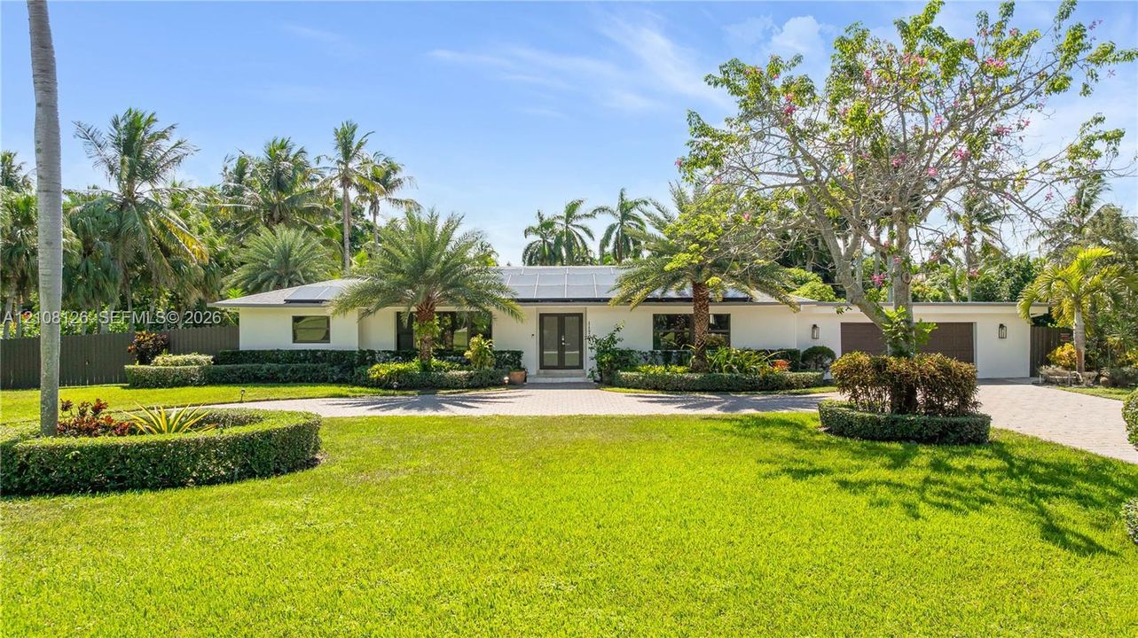 Single-story exterior featuring a light-toned stucco façade, solar panels, paver driveway, manicured landscaping, and mature palm trees