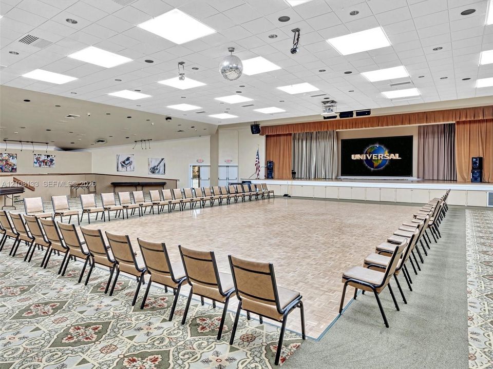 Expansive multi-purpose hall featuring a central parquet dance floor, a raised stage with built-in projection screen, tiered tray ceiling with recessed lighting, and integrated audio-visual equipment