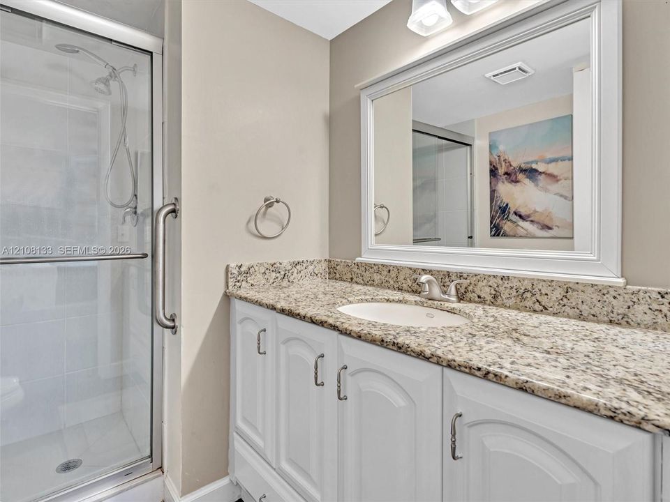 Bathroom vanity with stone-finish countertop, undermount sink, white cabinetry, wall-mounted mirror, and glass-enclosed shower