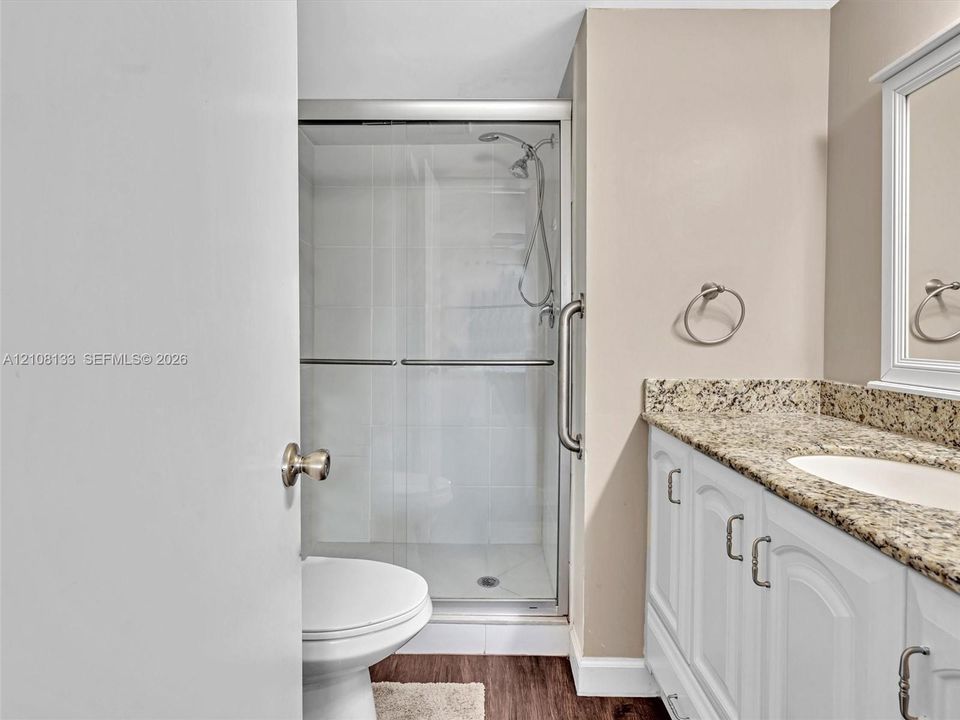 Bathroom featuring a vanity with stone-finish countertop, white cabinetry, a glass-enclosed shower with tile surround, and wood-finish flooring