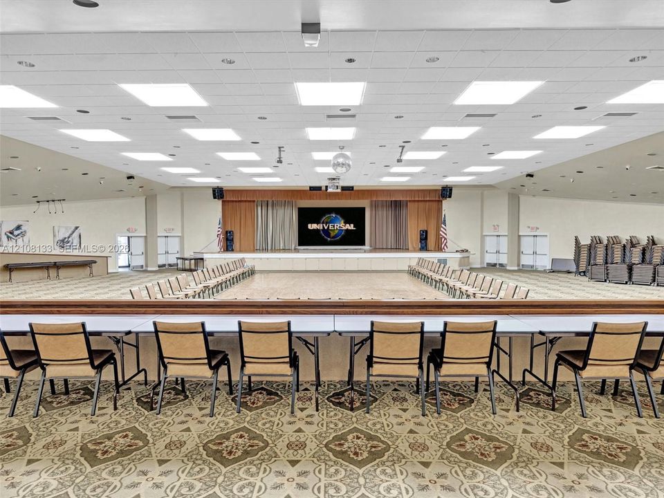 Large banquet hall featuring a recessed stage, wood-finish flooring, acoustic drop ceiling with integrated lighting, and patterned carpeting