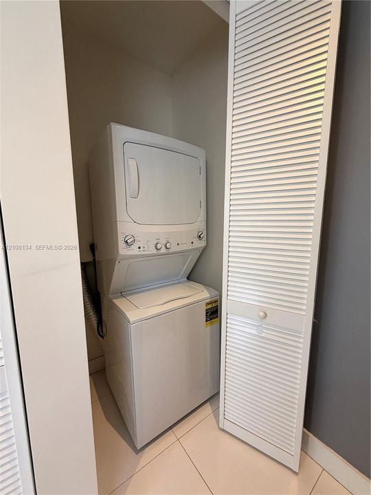 Stacked laundry unit with white finish, concealed within a closet featuring white louvered doors and neutral tile flooring