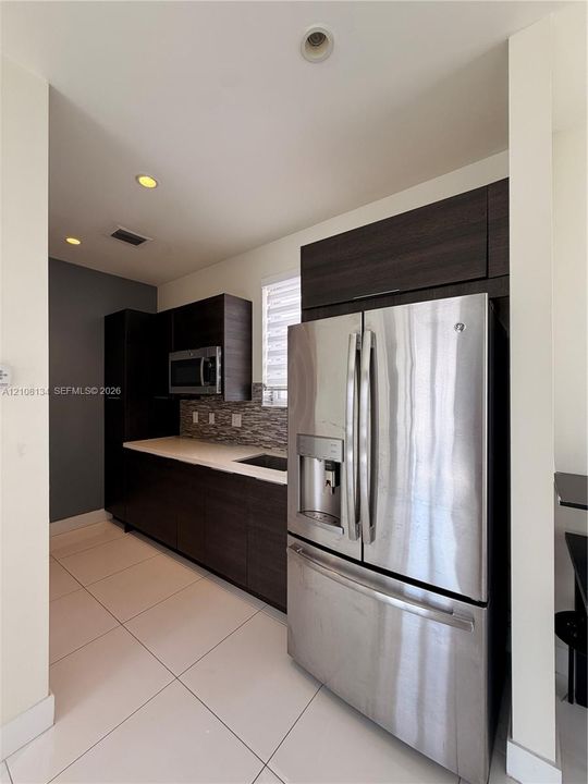 Kitchen area featuring dark wood-finish cabinetry, stainless steel French-door refrigerator, light-toned stone-finish countertops, mosaic tile backsplash, and large-format porcelain floor tiling