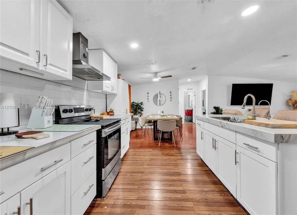 Kitchen featuring white cabinetry, wood-finish flooring, stone-surfaced island with integrated sink, stainless steel range with overhead hood, and white tile backsplash