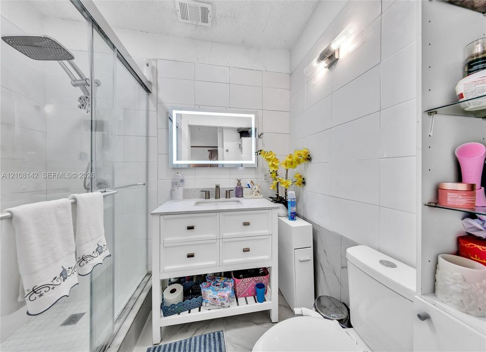 Bathroom featuring a walk-in shower with glass enclosure, white square tile wall surround, vanity with integrated basin, LED-backlit mirror, and built-in shelving