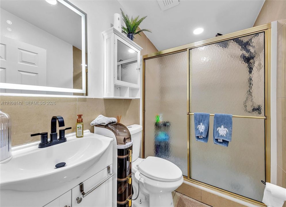 White vanity featuring a rectangular integrated sink, matte black widespread faucet, LED-backlit mirror, and walk-in shower with gold-tone sliding frame