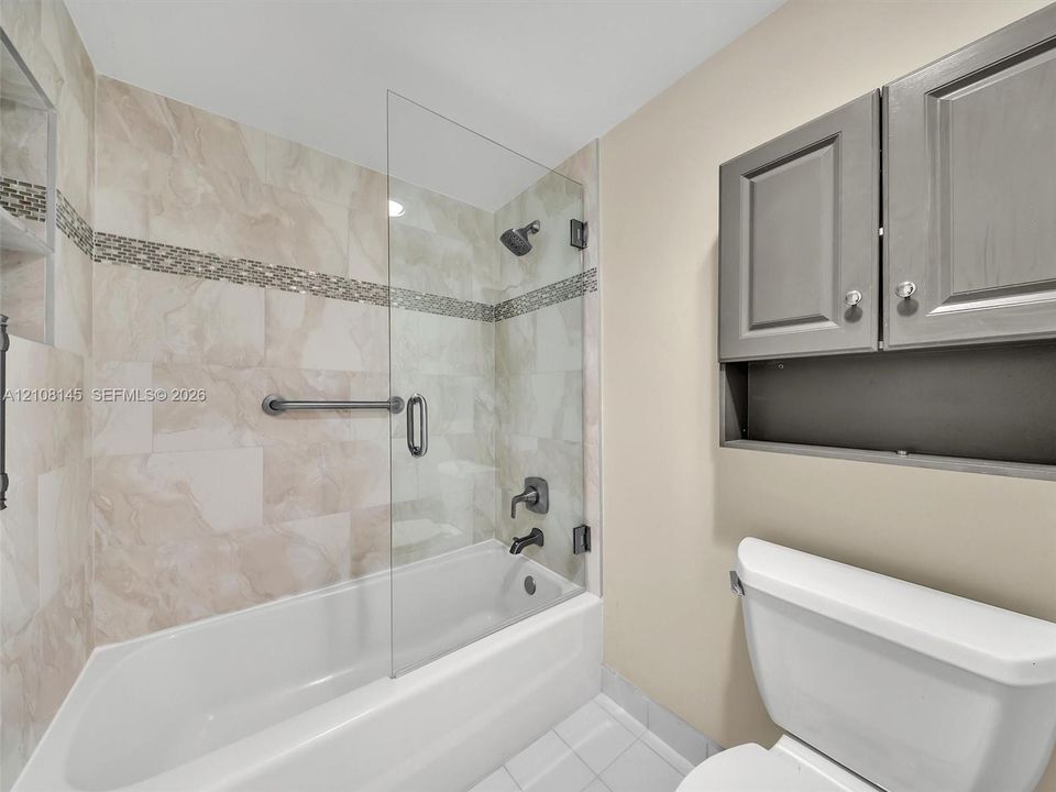 Bathtub featuring a glass shower panel, neutral tile surround with decorative mosaic accent, matte black wall-mounted fixtures, and built-in grey cabinetry