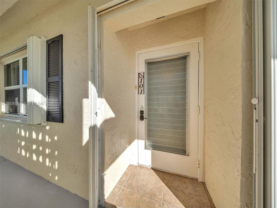Exterior entry featuring stucco finish, louvered shutters, tile flooring, and a glass-paneled storm door