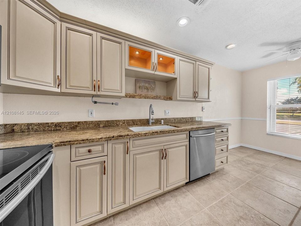 Kitchen featuring light-toned raised-panel cabinetry, stone-finish countertops, stainless steel dishwasher, integrated glass-front display shelving, and neutral tile flooring