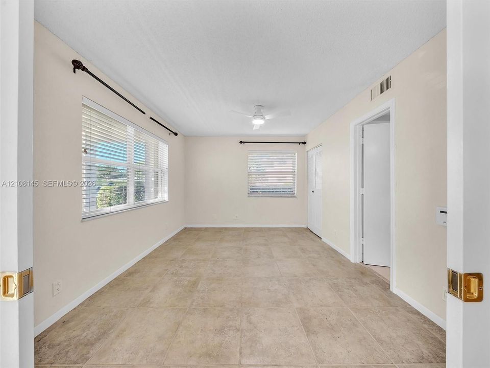 Neutral-toned room featuring large-format tile flooring, dual windows with horizontal blinds, a central ceiling fan with integrated lighting, and white interior doors