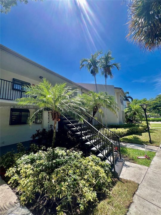 Exterior of multi-story building featuring stucco siding, black metal staircase railings, balcony, and mature palm landscaping