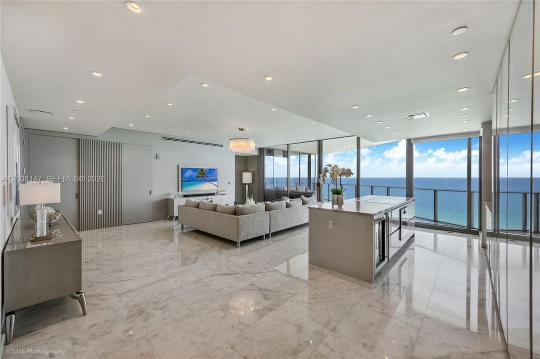 Open-concept living space featuring polished stone flooring, floor-to-ceiling glass wall panels, an integrated kitchen island with stone countertop, recessed ceiling lighting, and vertical-slat wall paneling