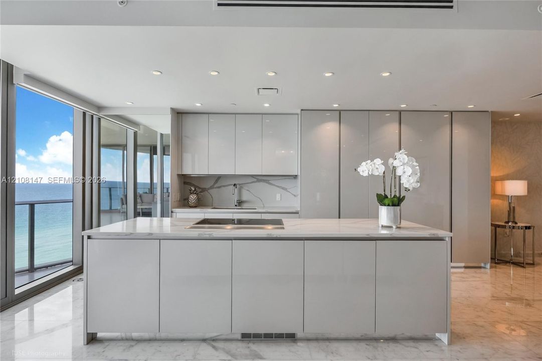 Kitchen featuring high-gloss cabinetry, stone-surface island with integrated cooktop, matching stone backsplash, polished tile flooring, and floor-to-ceiling windows