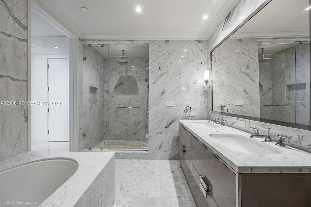 Bathroom featuring extensive stone-surround, frameless glass-enclosed shower with built-in recessed shelving, integrated soaking tub, custom dual-sink vanity with stone countertop, and wall-to-wall mirror
