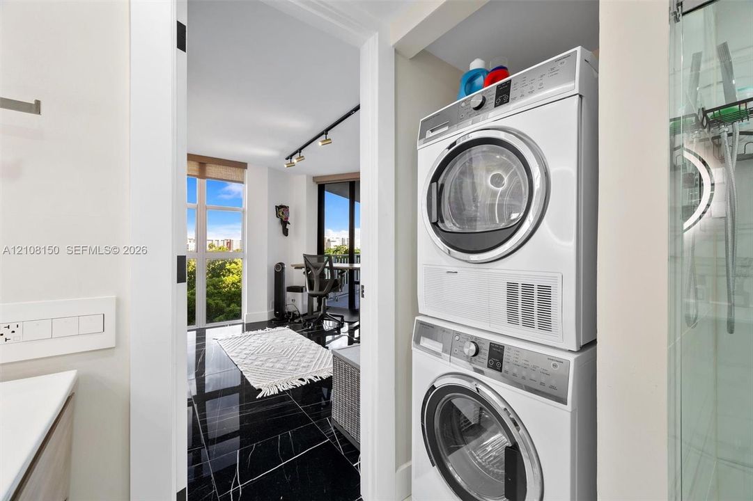 Stacked front-load laundry unit, black polished flooring with white veining, floor-to-ceiling windows, and track lighting