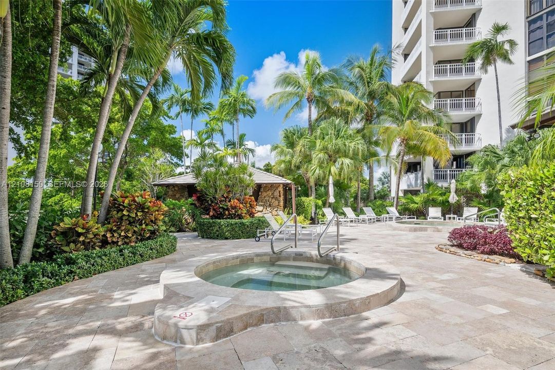 In-ground spa with stone surround, expansive stone-paved pool deck, mature landscaping, and multi-story building exterior