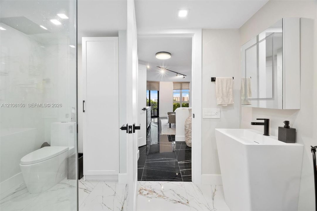 Modern bathroom featuring a tall white pedestal sink with matte black hardware, a glass-enclosed shower, built-in cabinetry, and white tile flooring with veining