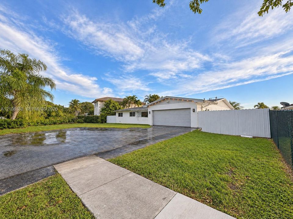 Exterior of a single-story residence featuring a two-car garage, expansive paved driveway, white privacy fencing, and a manicured lawn