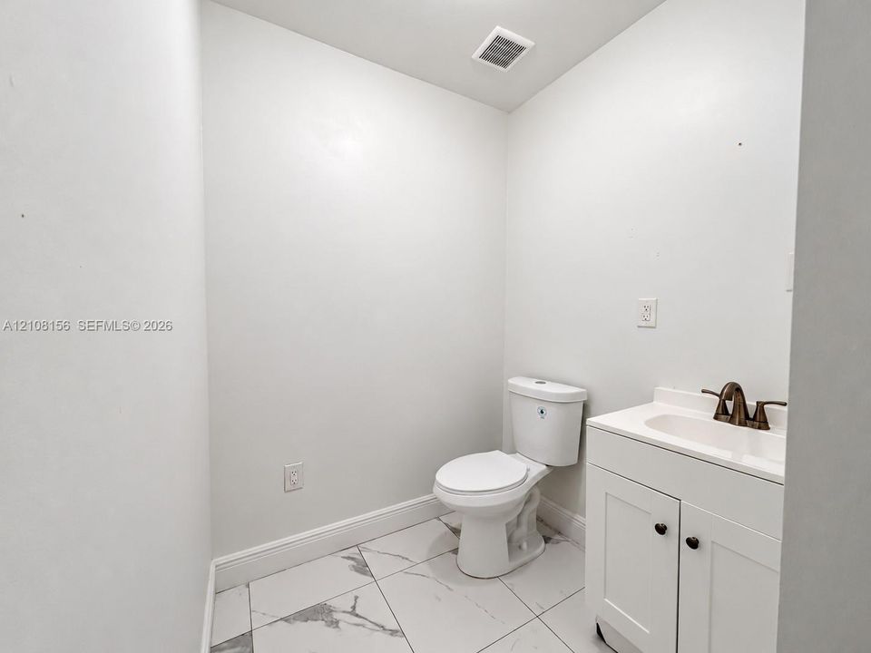 White vanity with a neutral countertop, dark metal faucet, ceramic toilet, and large-format tile flooring with a veined pattern