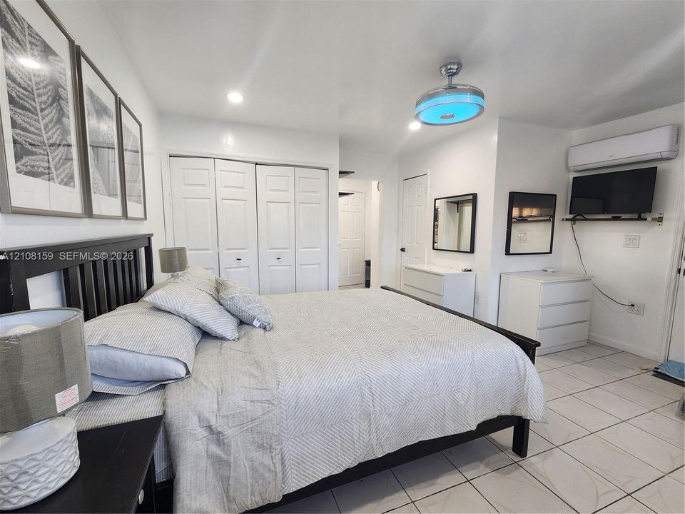 Neutral interior featuring white tile flooring, multi-panel sliding closet doors, ceiling-mounted light fixture with fan, and a wall-mounted ductless climate control unit