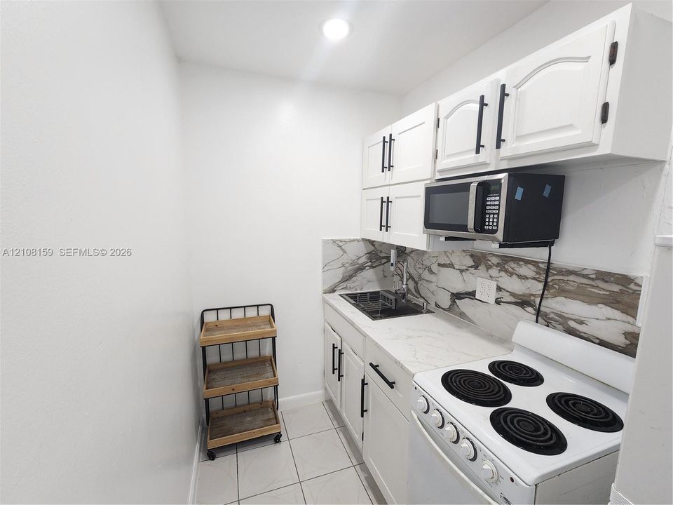 Kitchen area featuring white shaker-style cabinetry, stone-surface countertops, decorative stone-patterned backsplash, stainless steel sink, and integrated microwave