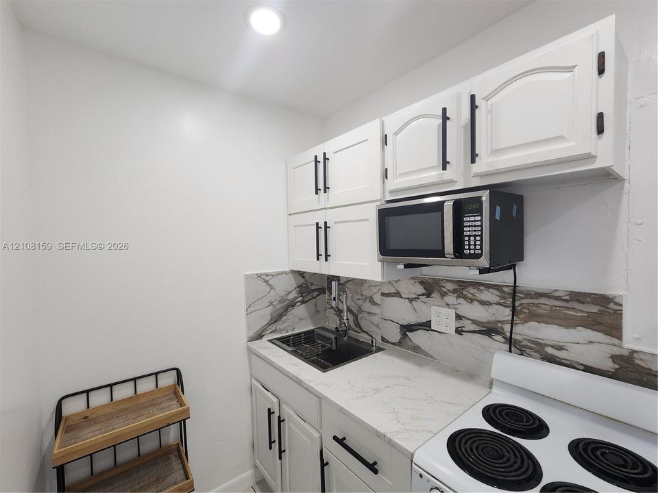 White shaker-style cabinetry with matte black hardware, stone-patterned backsplash, light-toned stone-finish countertop, stainless steel drop-in sink, and integrated microwave