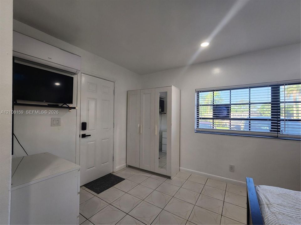 Room featuring neutral tile flooring, a wall-mounted climate control unit, a multi-pane window with horizontal blinds, and a built-in wardrobe with a mirrored door