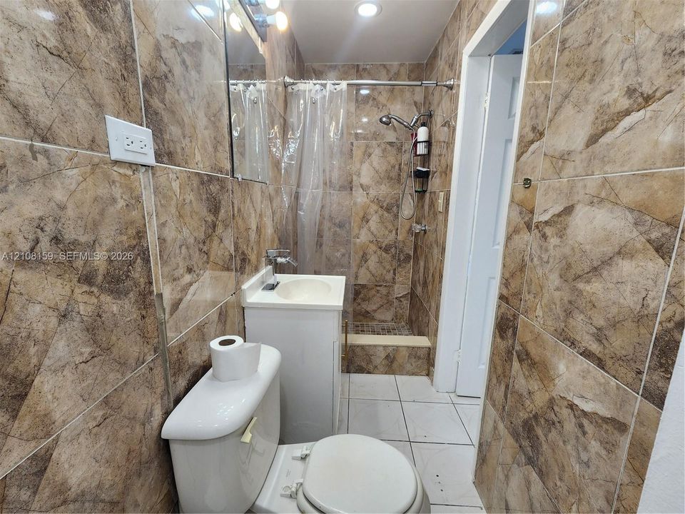 Bathroom featuring full-height stone-patterned wall tile, shower stall with a handheld fixture, pedestal sink with a chrome faucet, and white tile flooring