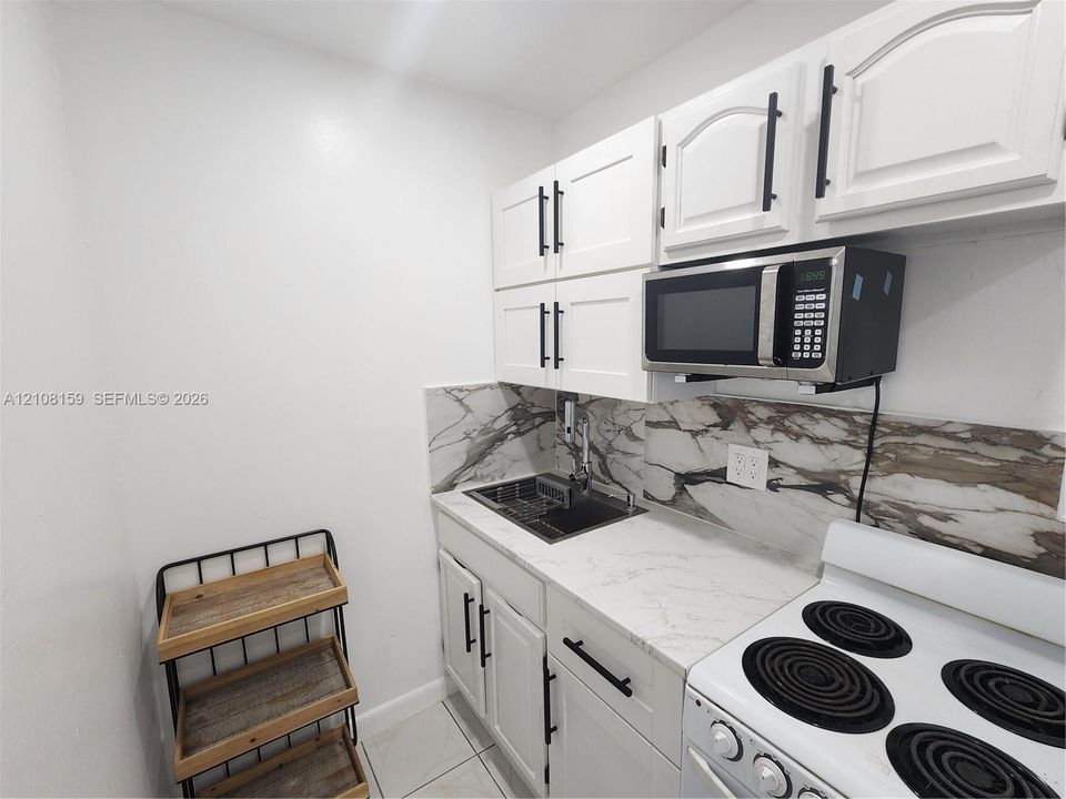 Kitchenette featuring white Shaker-style cabinetry, matte black hardware, stone-patterned backsplash, light-toned countertop, and integrated microwave