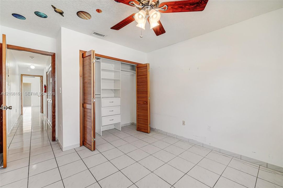 Ceramic tile flooring, built-in closet storage system with integrated shelving and drawers, wood-finish louvered bi-fold closet doors, and ceiling fan with integrated lighting