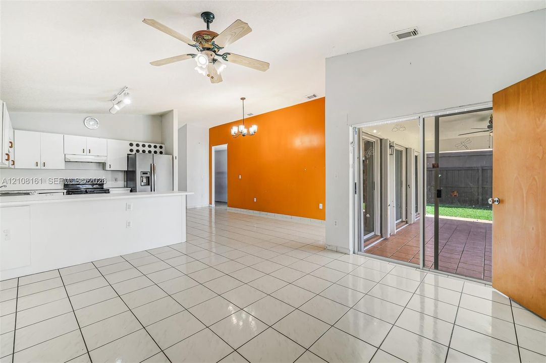 Open-concept area featuring white cabinetry, ceramic tile flooring, a ceiling fan with integrated lighting, a multi-arm chandelier, and sliding glass doors leading to a covered patio