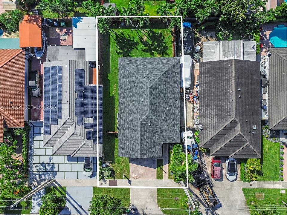 Aerial perspective of the property featuring a composition shingle roof, paved driveway, and grass lawn framed by perimeter fencing and mature palm trees