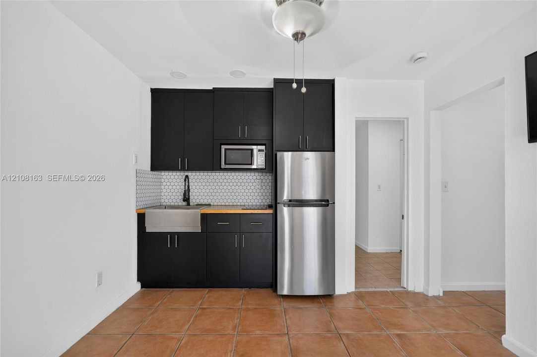 Kitchenette featuring dark flat-panel cabinetry, stainless steel farmhouse-style sink with matte black faucet, hexagonal tile backsplash, wood-tone countertop, and stainless steel refrigerator