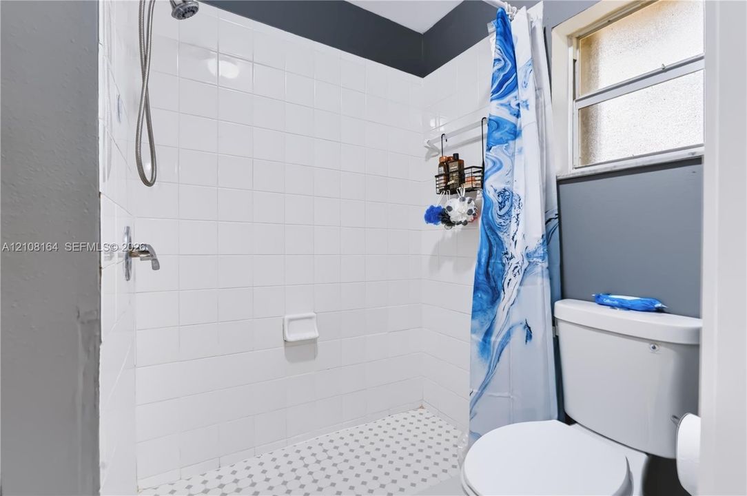 Full-height white tile shower enclosure with integrated soap dish, chrome shower hardware, patterned mosaic tile flooring, and standard-height toilet