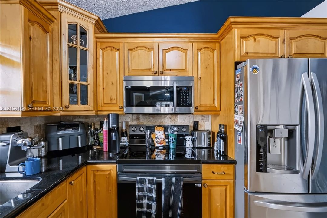 Kitchen featuring natural wood cabinetry, polished black stone countertops, stone tile backsplash, and stainless steel appliances including a built-in microwave and refrigerator
