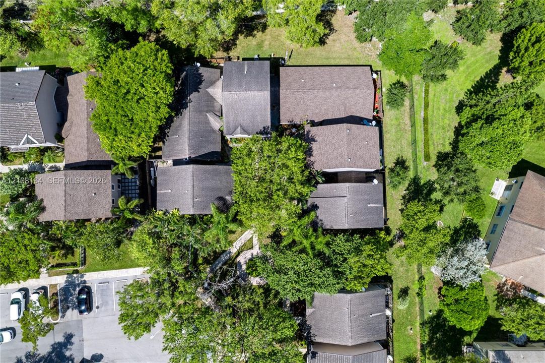Aerial view featuring a multi-unit property with shingled rooflines, surrounding mature trees, and ground-level paved parking