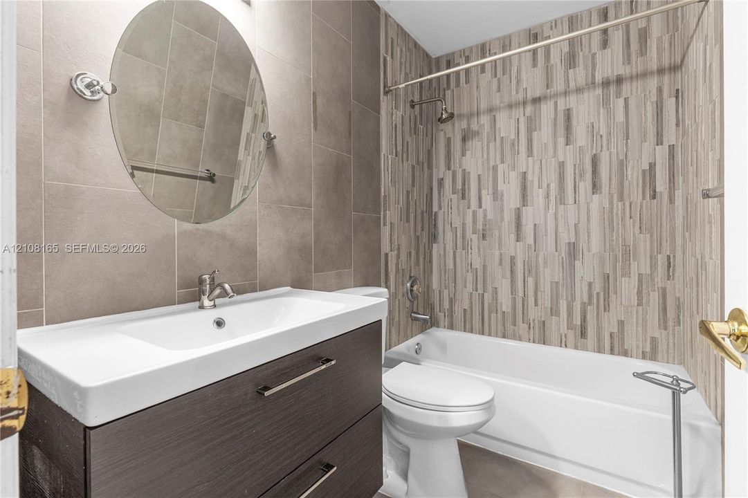 Bathroom featuring a wall-mounted vanity with a rectangular basin, large-format matte tile wall cladding, a decorative vertical-patterned tile shower surround, a circular mirror, and chrome fixtures