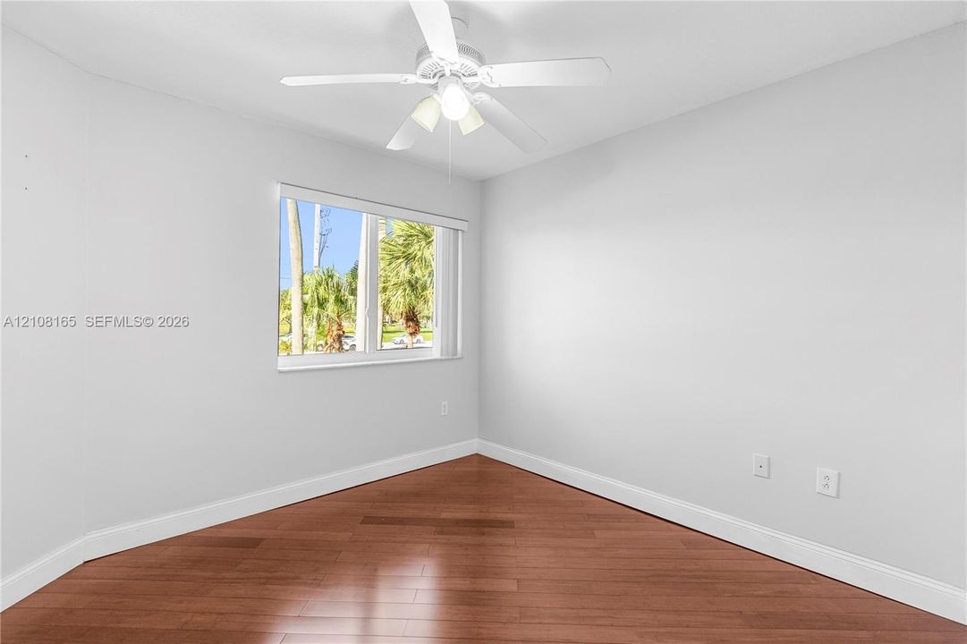 Neutral-toned interior room featuring wood-finish flooring, a white ceiling fan with integrated lighting, and a window with a built-in blind
