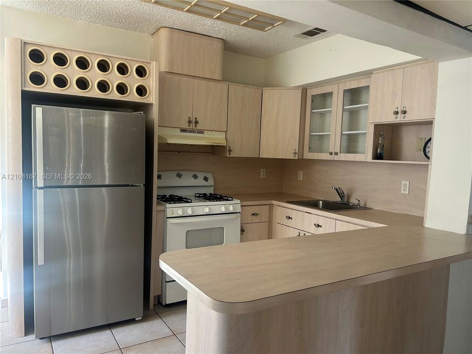 Kitchen featuring light wood-finish cabinetry, a peninsula countertop, stainless steel refrigerator, white gas range, and upper cabinets with glass-front paneling
