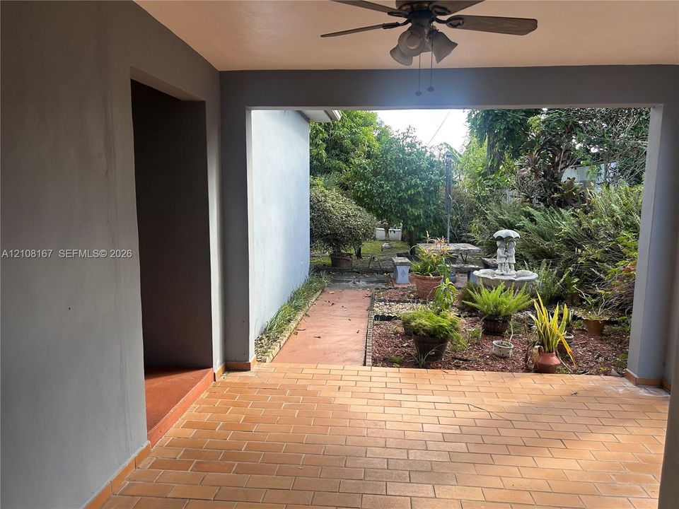 Covered patio featuring terra cotta tile flooring, a mounted ceiling fan, and access to a landscaped backyard with a stone-style fountain and established greenery