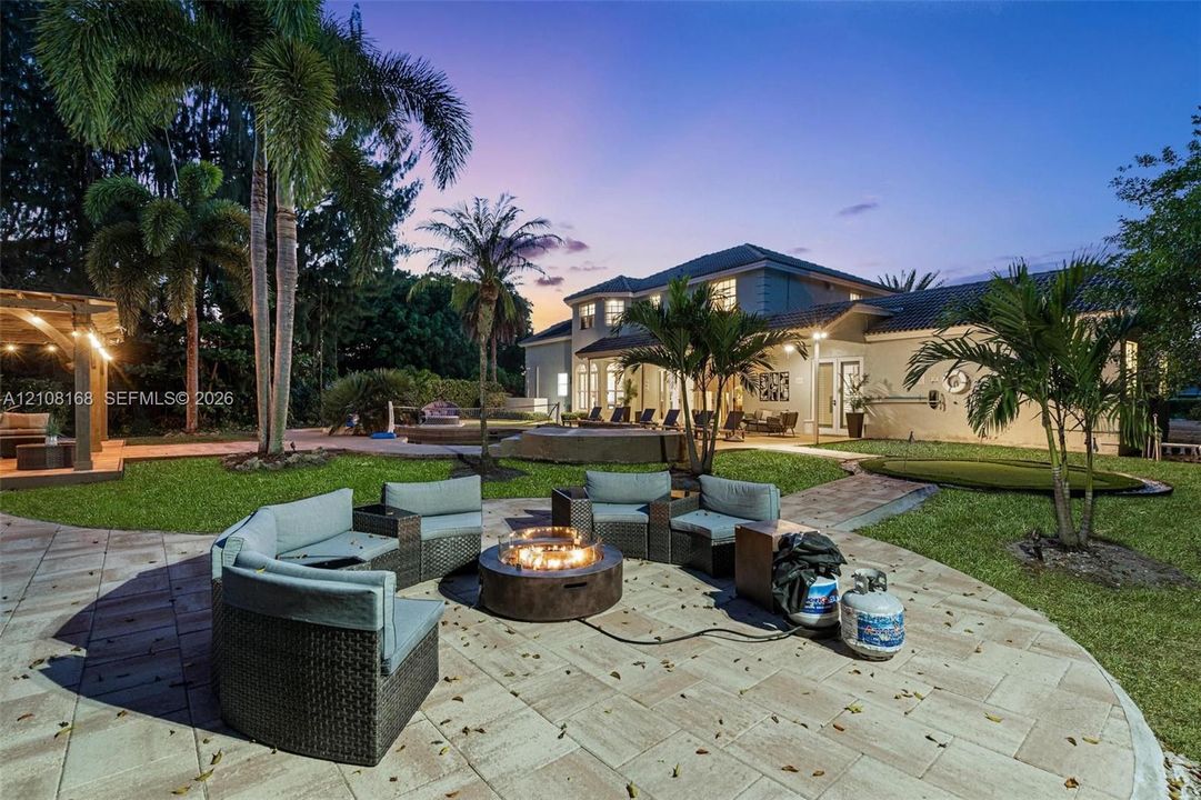 Expansive outdoor living area featuring paved hardscaping, a recessed swimming pool with spa, a wooden pergola, and mature palm trees