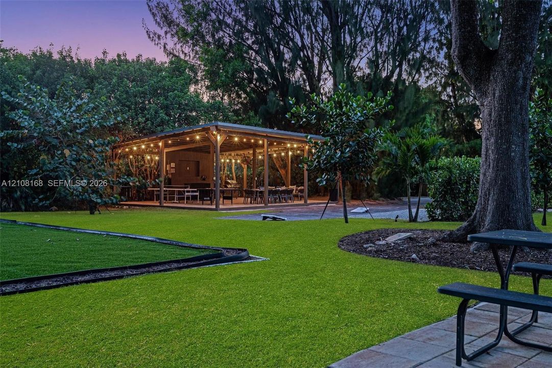 Expansive backyard featuring a covered wood-frame pavilion, manicured lawn, stone paver patio, and mature trees