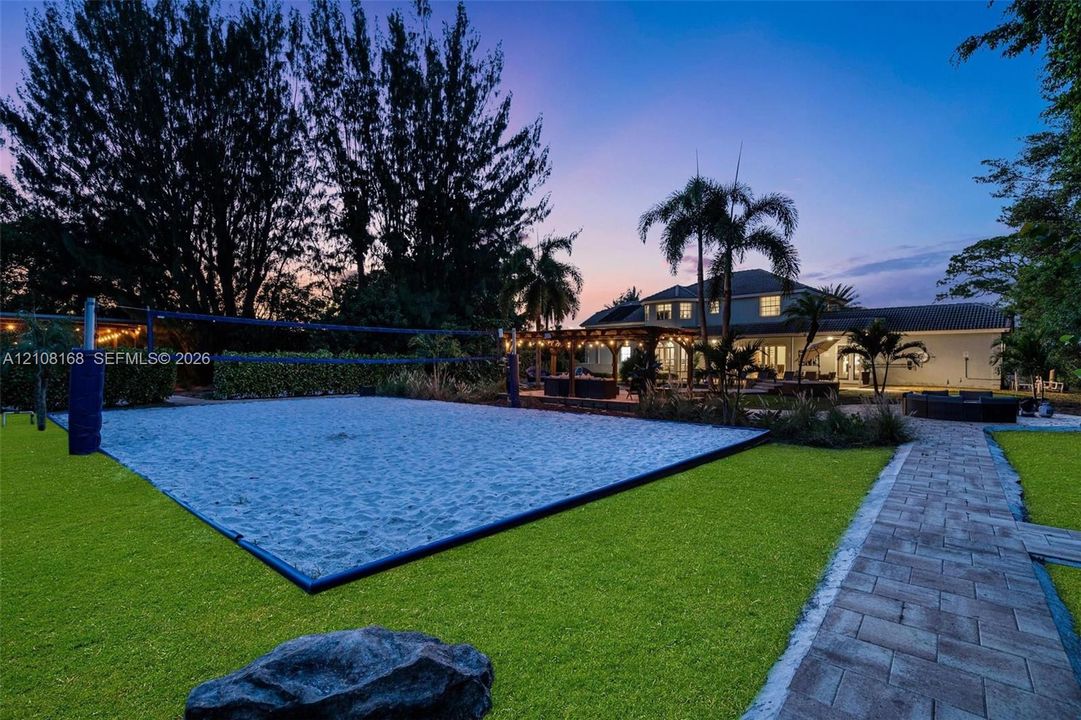 Sand volleyball court featuring a net and perimeter border, synthetic lawn, paver stone walkway, and covered outdoor structure with recessed lighting