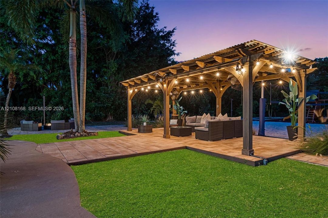 Backyard featuring a wood-frame pergola, stone-paver patio, manicured lawn, and mature palm trees