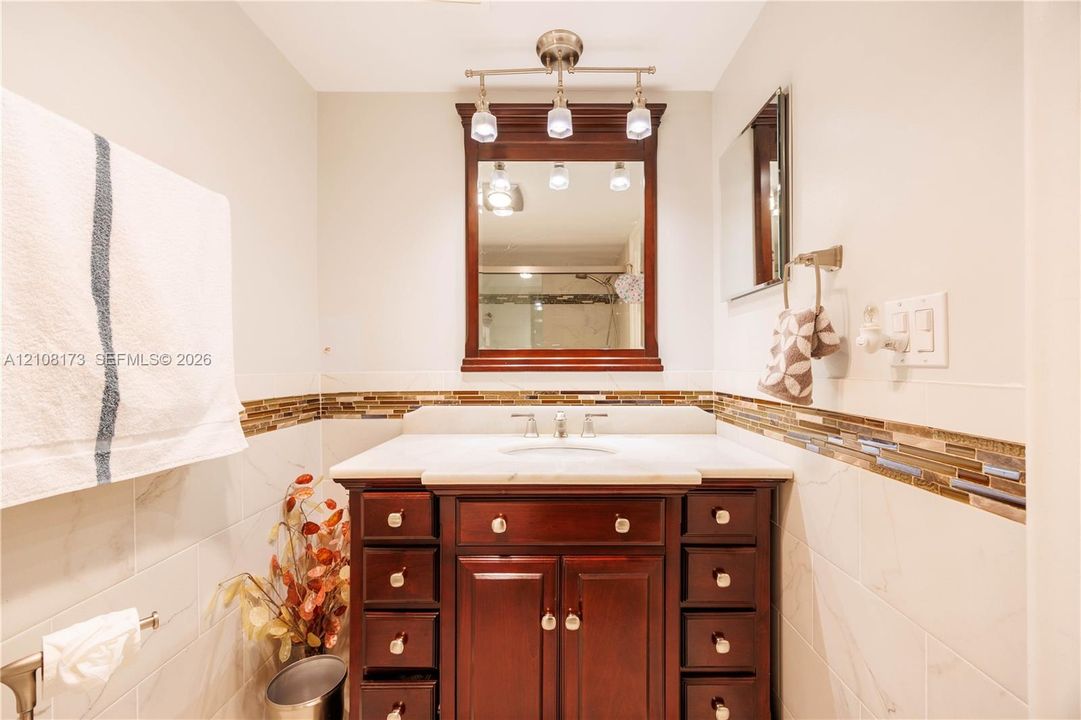 Bathroom vanity featuring wood cabinetry, stone countertop with an integrated basin, framed wall mirror, multi-bulb overhead lighting, and decorative glass tile backsplash