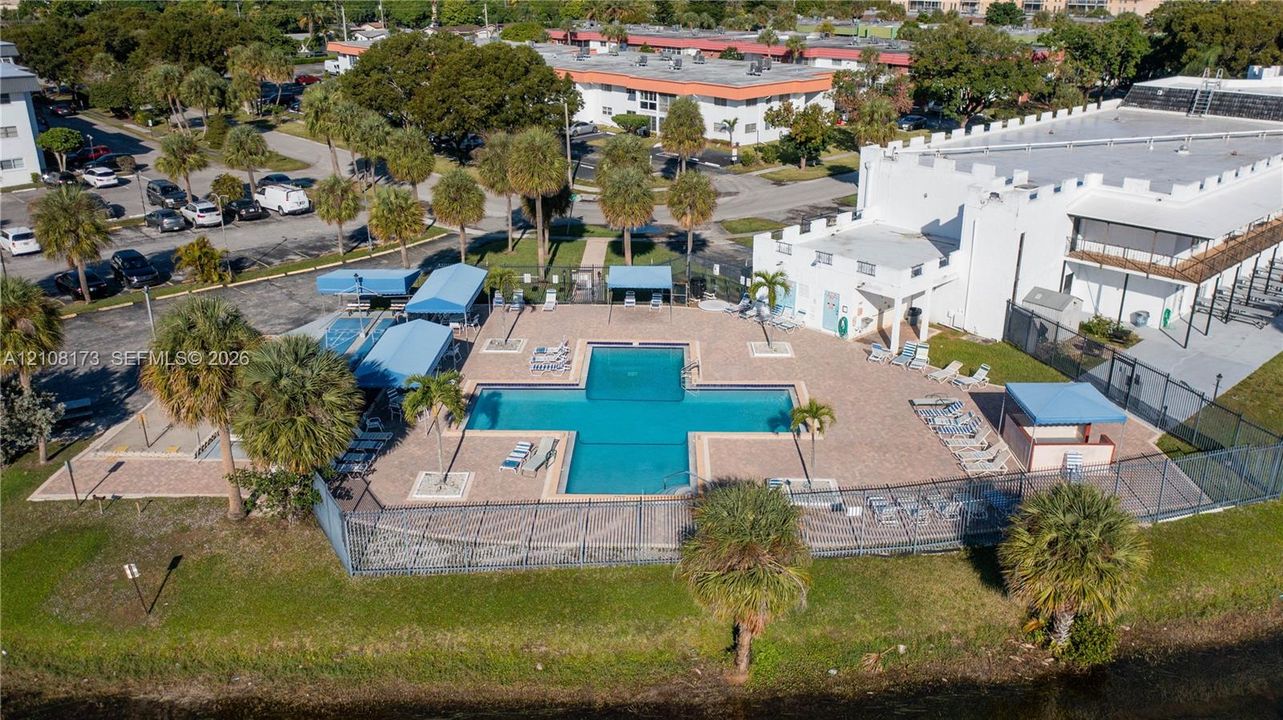 Outdoor pool with surrounding paver deck, permanent shade structures, mature palm trees, and metal perimeter fencing