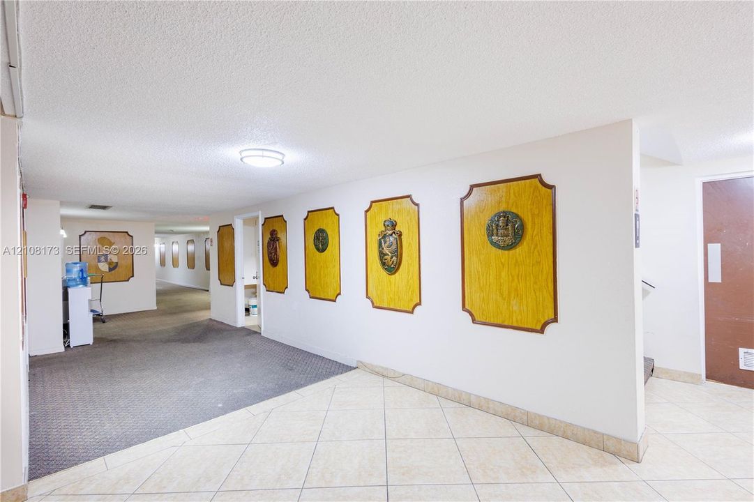 Interior common area featuring neutral-toned tile flooring, carpeted hallway, flush-mount ceiling lighting, and decorative wall-mounted plaques