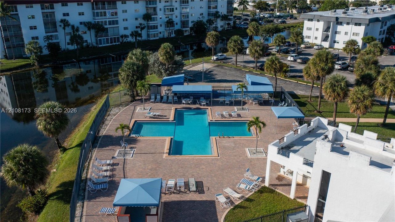 Community swimming pool featuring paver decking, multiple fabric shade structures, and surrounding landscaped grounds