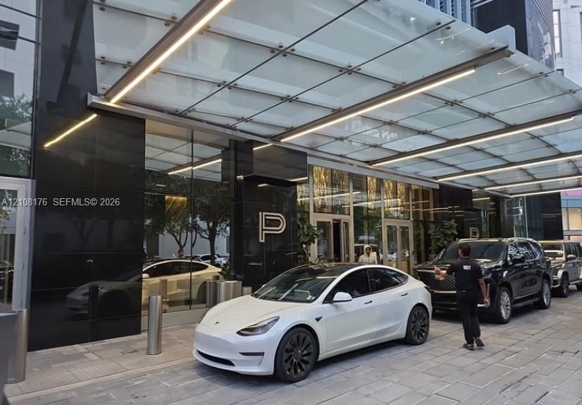 Covered porte-cochère featuring a glass canopy with integrated linear lighting, polished black stone facade, and stone-paved motor court