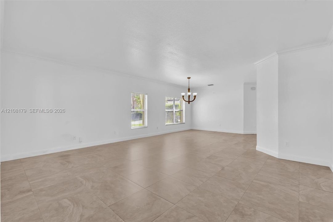 Open-concept interior featuring large-format tile flooring, recessed crown molding, dual windows, and a multi-arm chandelier